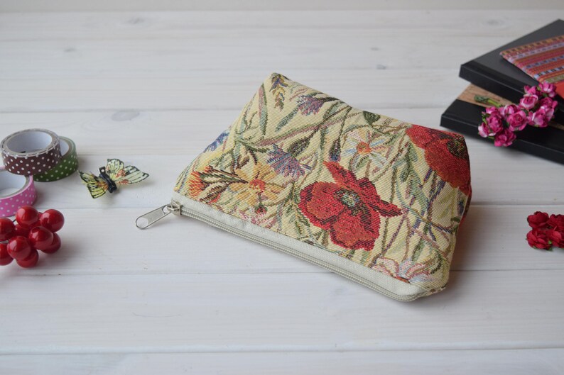 Pu&ograve; includere: Una pochette beige con cerniera con un motivo floreale che presenta papaveri rossi, fiori gialli e viola e foglie verdi. La pochette &egrave; realizzata in tessuto a tappezzeria.