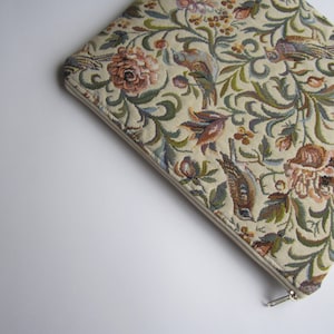 May include: A beige zippered pouch with a floral tapestry design featuring birds, flowers, and leaves in shades of green, brown, pink, and blue.