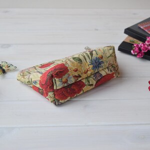 Pu&ograve; includere: Una piccola pochette rettangolare con cerniera, con un motivo floreale su sfondo crema e fiori rossi, gialli e verdi. La pochette &egrave; appoggiata su una superficie di legno bianco.