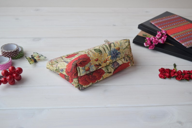 Pu&ograve; includere: Una piccola pochette rettangolare con cerniera, con un motivo floreale su sfondo crema e fiori rossi, gialli e verdi. La pochette &egrave; appoggiata su una superficie di legno bianco.
