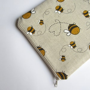 Pode incluir: Uma bolsa bege com zíper com um padrão repetitivo de abelhas amarelas e pretas voando. As abelhas são de estilo de desenho animado e têm um design simples.