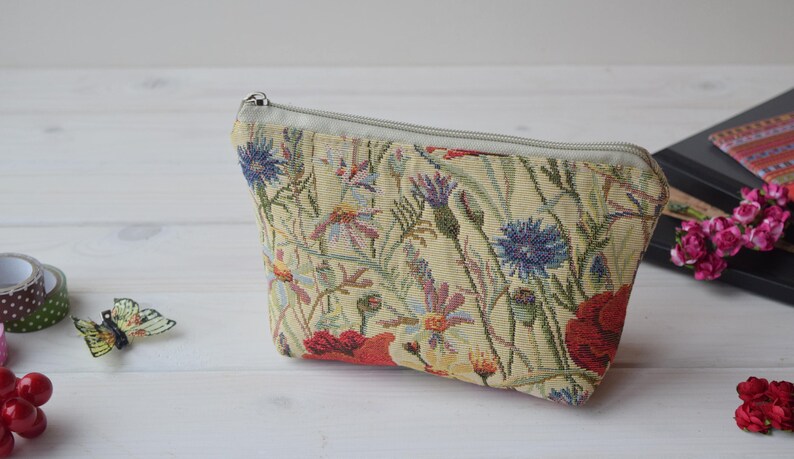 Pu&ograve; includere: Una pochette beige con cerniera con un motivo di arazzo floreale con papaveri rossi, fiordalisi blu e foglie verdi.