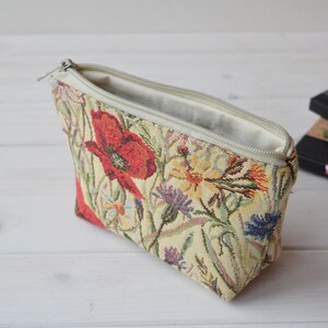 Pu&ograve; includere: Una pochette beige con cerniera con un motivo floreale con papaveri rossi, fiori gialli e foglie verdi. La pochette &egrave; aperta e la fodera &egrave; bianca.