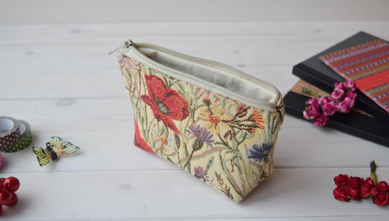 Pu&ograve; includere: Una pochette beige con cerniera con un motivo floreale con papaveri rossi, fiori gialli e foglie verdi. La pochette &egrave; aperta e la fodera &egrave; bianca.