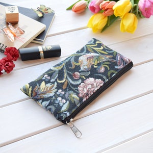 May include: A black zippered pouch with a floral tapestry design. The pouch is lying on a white wooden surface.