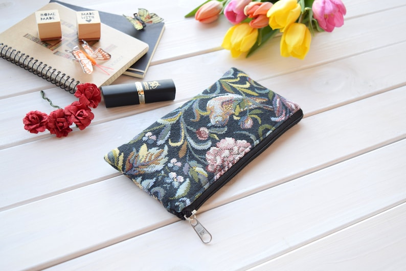 May include: A black zippered pouch with a floral tapestry design. The pouch is lying on a white wooden surface.