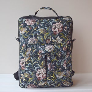 Peut inclure: Sac à dos en tapisserie florale avec un motif d'oiseaux et de fleurs dans des tons de rose, vert et crème sur fond bleu foncé. Il possède une poignée supérieure, des détails en cuir noir et des bretelles réglables.
