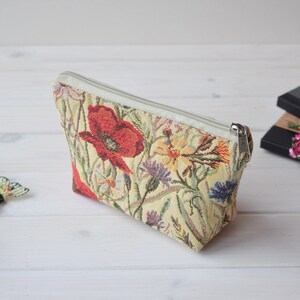 Pu&ograve; includere: Una pochette beige con cerniera con un design a tappezzeria floreale con papaveri rossi, fiori gialli e bianchi e foglie verdi.