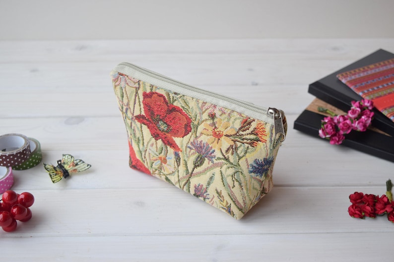 Pu&ograve; includere: Una pochette beige con cerniera con un design a tappezzeria floreale con papaveri rossi, fiori gialli e bianchi e foglie verdi.