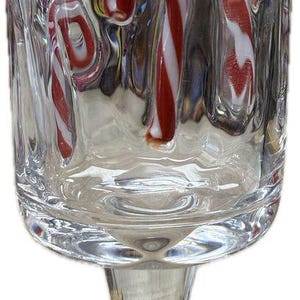 May include: A clear glass stemware piece featuring a textured surface with embedded red and white candy cane motifs. The glass has a cylindrical bowl and a long, slender stem, showcasing a festive design with distinct striped patterns.