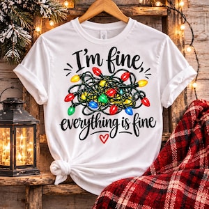 May include: A white short-sleeve t-shirt featuring a graphic of a tangled mess of colourful Christmas lights in red, green, yellow, and blue. The text reads I'm fine, everything is fine in black script, with a small red heart icon below.