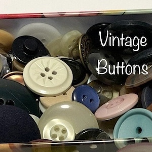 May include: A tin box filled with a variety of vintage buttons in different colours, shapes, and sizes. The buttons are made of plastic, metal, and fabric. The box is labelled "Vintage Buttons".