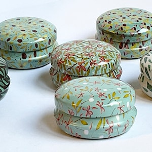 May include: Six round, stackable, metal tins with different floral and leaf patterns. The tins are in shades of green, brown, and white.