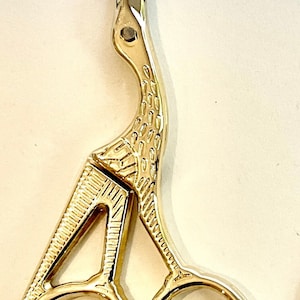 May include: Gold-coloured, decorative scissors with a bird-shaped handle. The scissors have two rings for the fingers and a pointed tip.