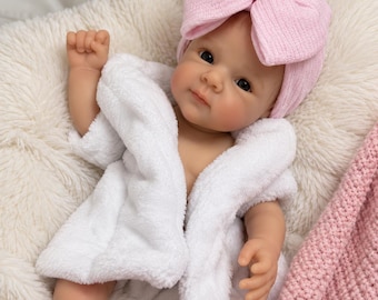 May include: A realistic baby doll with light skin, dark eyes, and a neutral expression. The doll wears a soft white terry cloth robe and a light pink ribbed knit turban-style hat with a central bow detail, resting on a textured surface.