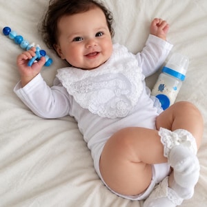 May include: A baby wearing a white long-sleeve bodysuit, a white lace-trimmed bib, and white lace-trimmed socks. The baby holds a blue and white beaded rattle and is positioned next to a baby bottle with a blue cap and a blue base.
