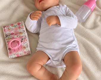 May include: A sleeping baby doll with light skin wearing a plain white long-sleeved bodysuit. Beside the doll is a pink plastic baby bottle and a floral-patterned fabric accessory featuring a pink pacifier attached to the front.