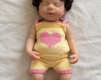 May include: A realistic baby doll with dark curly hair and a large pink bow. The doll wears a yellow knitted tank top with a pink heart design, matching yellow shorts with pink trim, and white slip-on shoes with small colorful patterns.