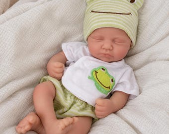 May include: A newborn doll with closed eyes wearing a light green and white striped beanie with frog features, a white short-sleeved shirt with a green frog applique, and matching light green and white striped shorts, resting on a white textured fabric.