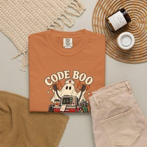 May include: A burnt orange t-shirt featuring a graphic of a ghost wearing a nurse cap, holding defibrillator paddles, and standing behind a medical cart with a heart monitor displaying the text CODE BOO in a retro font.