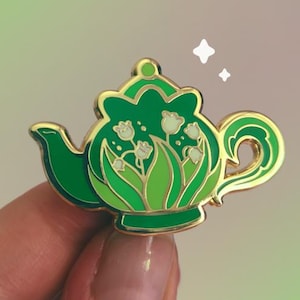May include: A gold-rimmed enamel pin shaped like a teapot in shades of green. The teapot features a central design of white lily of the valley flowers with green leaves. The pin has a glossy finish and includes a small white sparkle graphic above it.