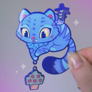 Peut inclure: Une broche en émail représentant un chat bleu avec un petit oiseau à chapeau sur le dos et une breloque cupcake suspendue à sa patte. Le chat a de grands yeux jaunes, des rayures bleu foncé et des touches roses sur les oreilles et les joues.