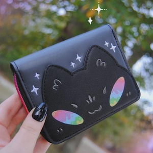 May include: A black bi-fold wallet featuring an embroidered cat face with iridescent rainbow-coloured cheeks and stitched whiskers. The wallet is decorated with small white embroidered stars and includes a bright pink zip along the side edge.