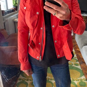 May include: A bright red jacket with a stand-up collar, multiple pockets, and silver-toned hardware. The jacket has a zipper closure and snap buttons. The person is wearing blue jeans.