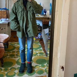 Può includere: Una persona indossa una giacca verde in stile militare con chiusura a zip e bottoni, jeans blu e stivaletti neri. La persona è in piedi su un tappeto a motivi con cerchi verdi e gialli.
