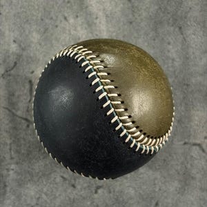 Puede incluir: Una pelota de béisbol con un diseño bicolor, con una mitad en negro mate y la otra en verde oliva apagado. Ambas secciones están unidas por una costura prominente y elevada, realizada con hilo grueso de color claro en diagonal.