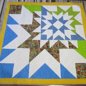 May include: A colorful quilt with a starburst design. The quilt features a central star pattern in blue and white, surrounded by green, white, and patterned fabric with cacti and floral designs. The quilt is bordered with a yellow edge.
