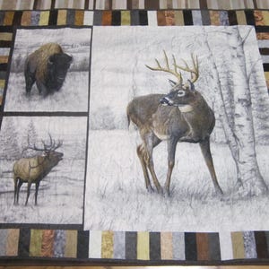 Puede incluir: Una colcha acolchada con escenas de fauna. Presenta un bisonte, un ciervo con cuernos y un alce, todos sobre un fondo nevado. El borde es un patchwork de varios colores y patrones.