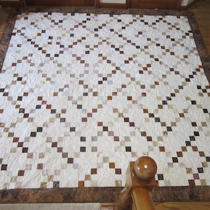 May include: A rectangular quilt featuring a white background with a repeating diagonal grid pattern of small squares in various shades of brown, tan, and cream. The quilt is finished with a wide, solid dark brown border along all four edges.