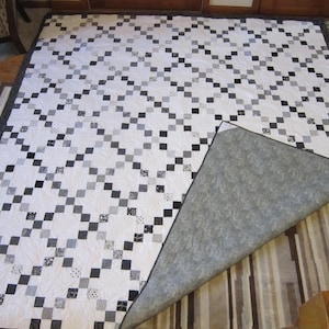 May include: A handmade quilt with a white background and a repeating pattern of black, gray, and light gray squares. The quilt's border is a dark gray, and the backing is a solid gray fabric. The quilt is displayed on a wooden surface.
