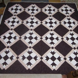 May include: A brown and white quilt with a geometric pattern. The quilt features a repeating diamond design with alternating white and brown squares, framed by a light tan border. The quilt is displayed on a wooden table.