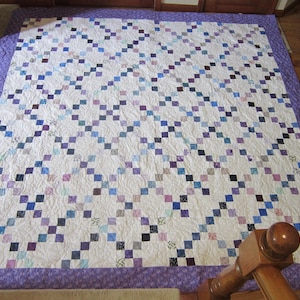 May include: A square quilt featuring a white background with a repeating diagonal grid pattern of small multi-colored squares in shades of blue, purple, and black. The quilt is finished with a wide purple border featuring a subtle floral print.