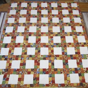 Puede incluir: Un edredón hecho a mano con un diseño de patchwork. El edredón presenta un patrón de cuadrícula de cuadrados blancos alternados y cuadrados de varios colores, incluyendo rojo, verde y dorado. El edredón está extendido sobre un suelo de madera.