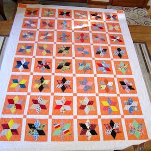 以下が含まれることがあります： オレンジ色の正方形と星型のパッチワークデザインが繰り返される手作りのキルト。星は赤、青、黄色、黒など、さまざまなカラフルな生地で作られています。キルトには白い縁取りがあります。