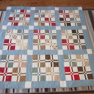May include: A handmade quilt with a patchwork design. The quilt features a light blue border and a pattern of squares in various colors, including red, brown, and cream. The quilt is placed on a wooden table.