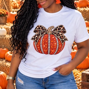 May include: A white short-sleeved t-shirt featuring a graphic of an orange pumpkin adorned with a leopard print bow. The pumpkin design is embellished with sparkling rhinestones and studs, creating a textured, decorative appearance.
