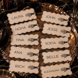 Puede incluir: Un conjunto de diez tarjetas de sitio rectangulares con bordes festoneados, cada una con un gráfico de mano de esqueleto negro y un nombre único: Laura, John, Noha, Jesse, Tina, Sofia, Ana, Belle, Rich y Alexandra, en fuente negra.