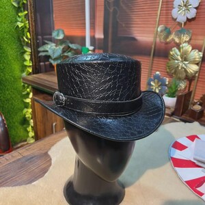 May include: A black top hat featuring a textured, embossed crocodile-skin pattern across the crown and brim. A smooth black band encircles the base of the crown, secured by a metallic silver-toned buckle detail on the left side of the hat.