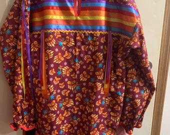 Puede incluir: Una camisa roja de manga larga con un estampado de huellas de patas de oso. La camisa tiene cuello en V y está decorada con ribetes de cinta de colores. La camisa se combina con pantalones negros con ribetes de cinta de colores.