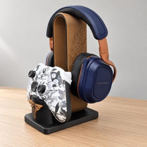 May include: A gaming headset and controller stand featuring a wooden arch with a fern leaf engraving. A dark blue Turtle Beach headset rests on the arch, while a white and gray patterned Xbox controller with a firefly emblem sits in front.
