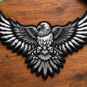 May include: An embroidered patch featuring a bald eagle with wings fully spread in flight. The design uses a monochromatic color palette of black, white, and shades of gray, with a thick black border outlining the entire eagle shape.