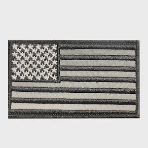 May include: A rectangular embroidered patch featuring the United States flag in a monochromatic black and grey color scheme. The design includes a field of stars on the left and horizontal stripes, all rendered with detailed stitching.