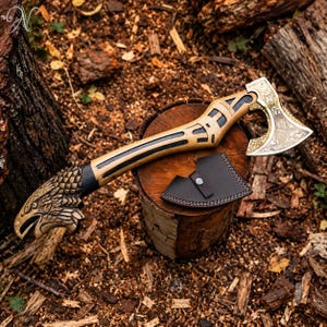 May include: A hand axe with a light wood handle featuring black accents and a carved eagle head pommel. The metal axe head has an engraved floral pattern and a hammered texture. A black leather sheath with a silver snap button is included.
