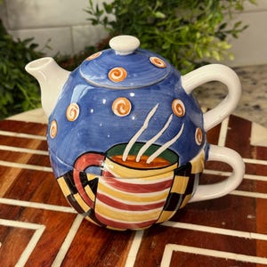 May include: A ceramic tea-for-one set featuring a blue teapot with orange spiral accents and a matching cup decorated with red, yellow, and white horizontal stripes and a black and yellow checkered pattern, stacked together as a single unit.