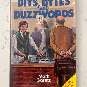 May include: A book titled Bits, Bytes and Buzzwords by Mark Garetz. The cover features an illustration of a person in a vest behind a counter with a computer, and another person in a brown jacket holding a briefcase. A yellow corner banner reads As Seen On The Popular TV Show Of The Same Name.