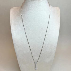 May include: A silver-toned necklace featuring a delicate chain with small, evenly spaced metallic links and a slender, vertical pendant encrusted with tiny, clear, sparkling stones that catch the light, hanging centered on a display bust.